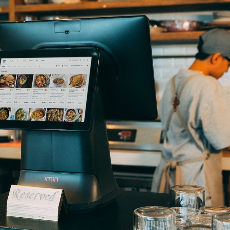 Digital ordering system in a restaurant kitchen with a touchscreen display and a chef at work.